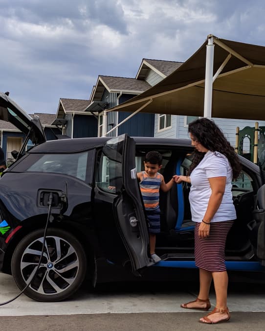 A person with long black wavy, white T-shirt, striped skirt and sandals holds the hand of a small child standing inside a Black electric car that has a charger cord plugged into its side.