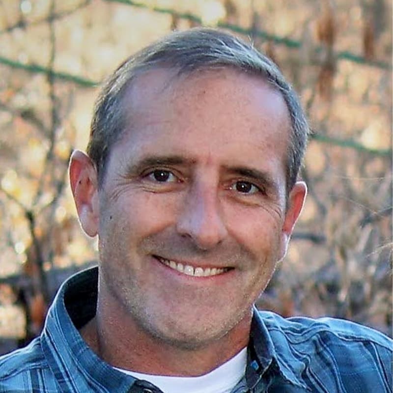 Headshot of Benj Fredrick, who is smiling and stands in front of tree branches. He has short gray hair and wears a blue button up tshirt.