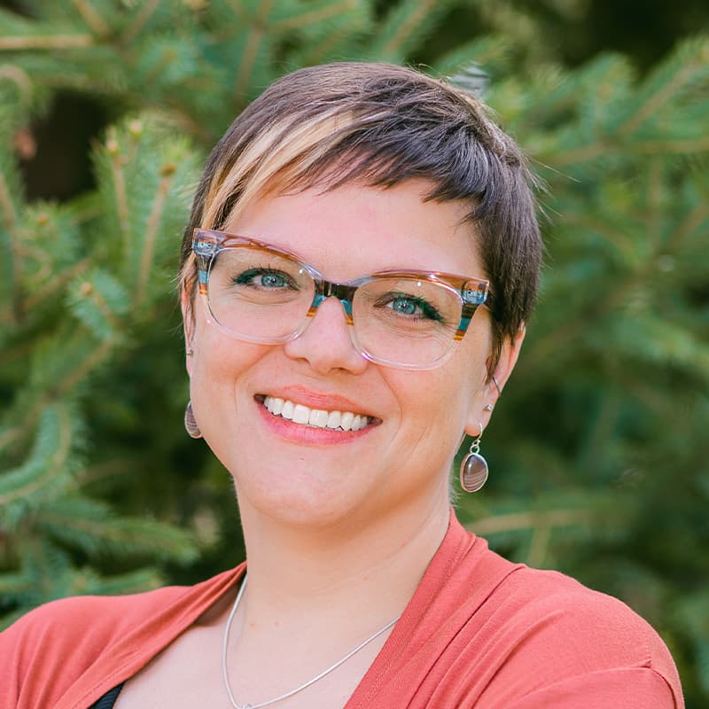 Headshot of Jenna Warmuth, who is smiling and has short light brown hair. She wears glasses and wears a salmon colored cardigan.