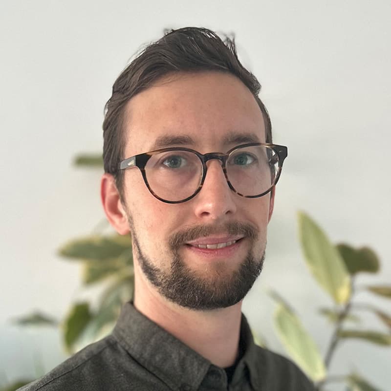 Headshot of Spenser Garden, who is slightly smiling and wears glasses. He has short brown hair and short facial hair.