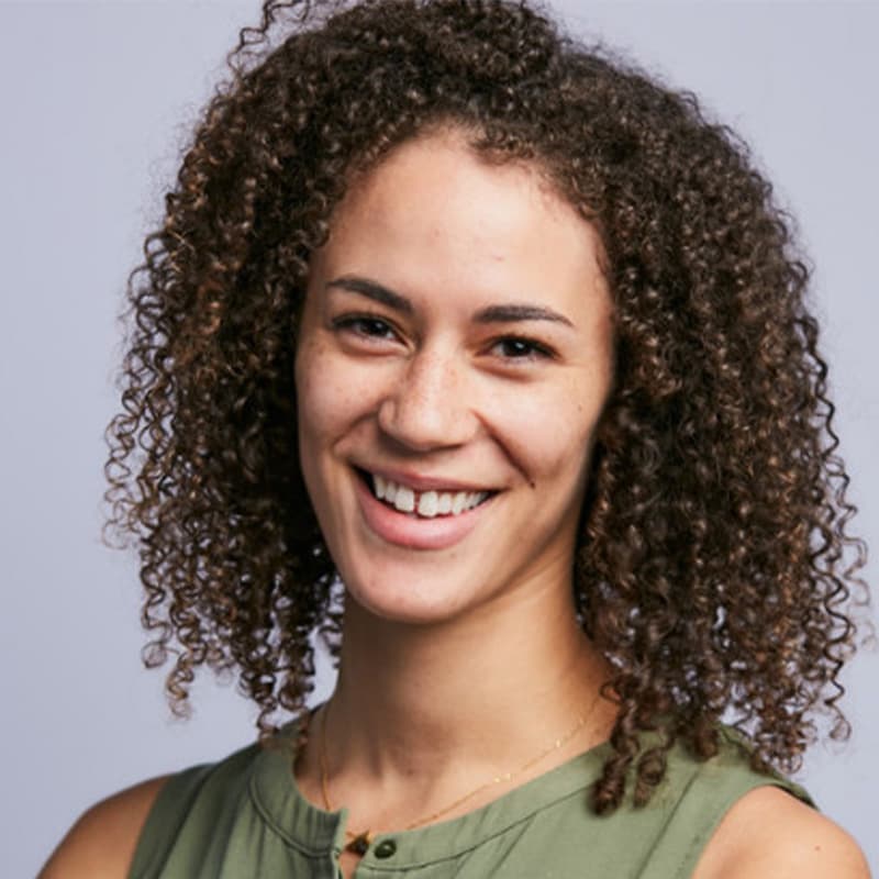 Headshot of Hilary Thompson, who is smiling at the camera and has brown curly hair. She wears a green sleeveless top with a gold necklace.