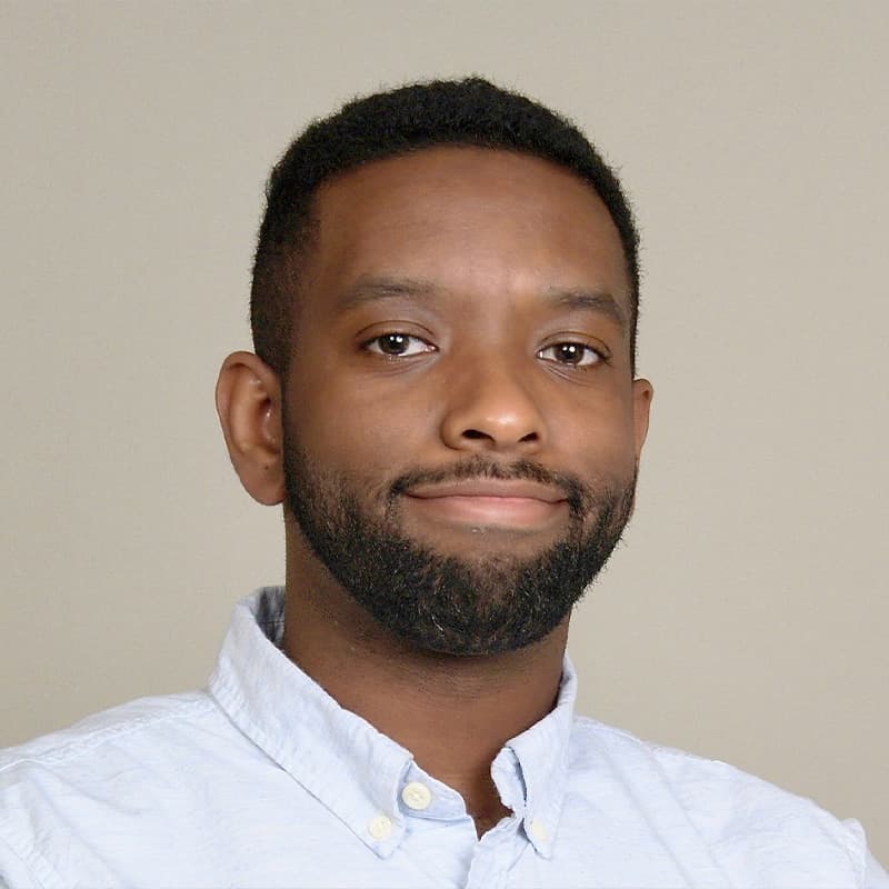 Headshot of Cameron Johnson-Brown, who is slightly smiling and has short black hair and short facial hair. He wears a light blue button up shirt.