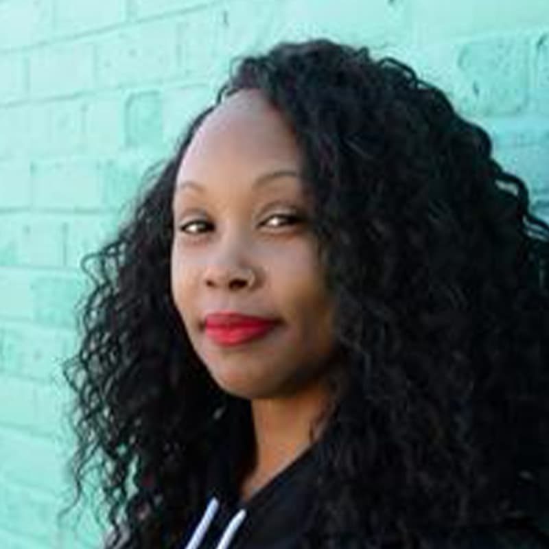 Headshot of Melissa Jones, who is smiling and has long curly black hair. She is wearing red lipstick and stands against a teal brick wall.