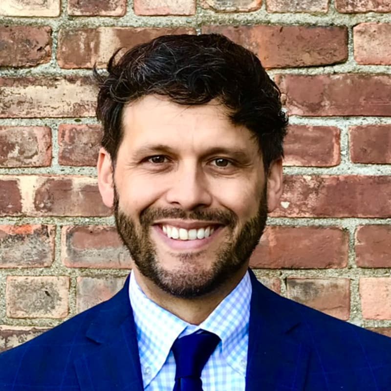 Headshot of Michael Hernandez, who is smiling and has short curly brown hair and short facial hair. He wears a navy suit and tie and is standing in front of a brick wall.