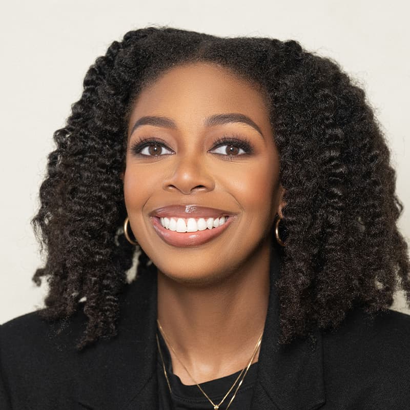 Headshot of Jessica Dulaney, who is smiling and has black curly hair. She is wearing a black suit and wears gold necklaces.