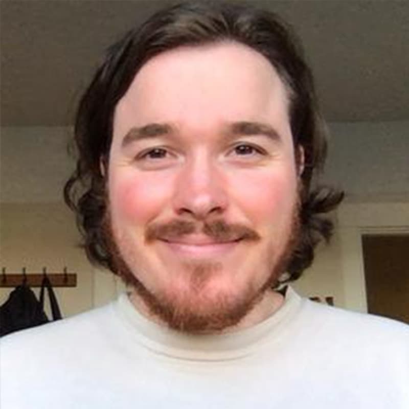 Headshot of Luke Donahue, who is smiling and has shoulder length brown curly hair and short facial hair. He is wearing a white crewneck.