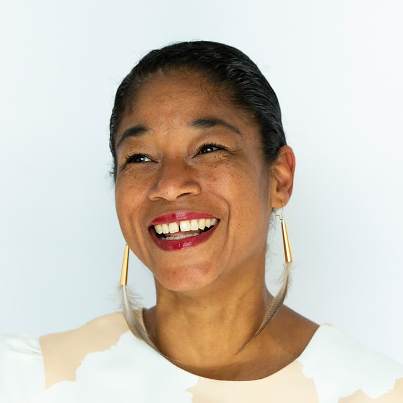 Headshot of Natalie Bomberry, who is smiling and has black hair pulled back into a ponytail. She wears feather earrings, red lipstick, and a beige top.