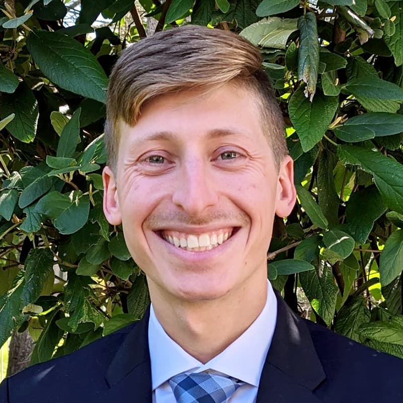 Headshot of Joshua Gilstein, who is smiling and has short blond hair. He is wearing a suit with a blue checkered tie.