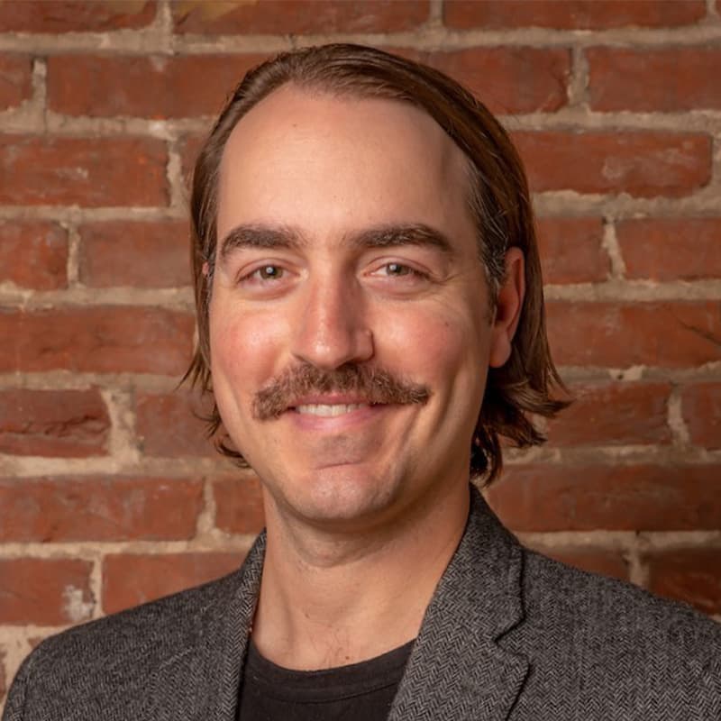 Headshot of Alex Amend, who has chin length brown hair and a mustache. He wears a gray jacket and stands in front of a brick wall.