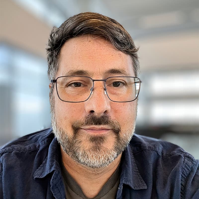 Headshot of Adam Roessler, who is slightly smiling and wears thin framed glasses. He has short brown hair and short facial hair, and wears a navy button up shirt.