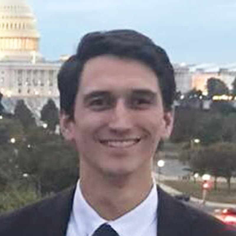 Headshot of Zach Pierce, who is smiling and wears a black suit. He has short black hair.